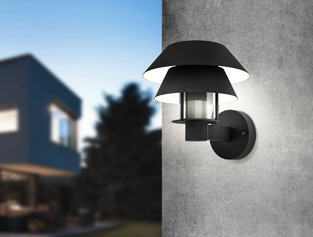 Mise en ambiance de l'applique noire allumée sur un mur gris, maison en arrière-plan