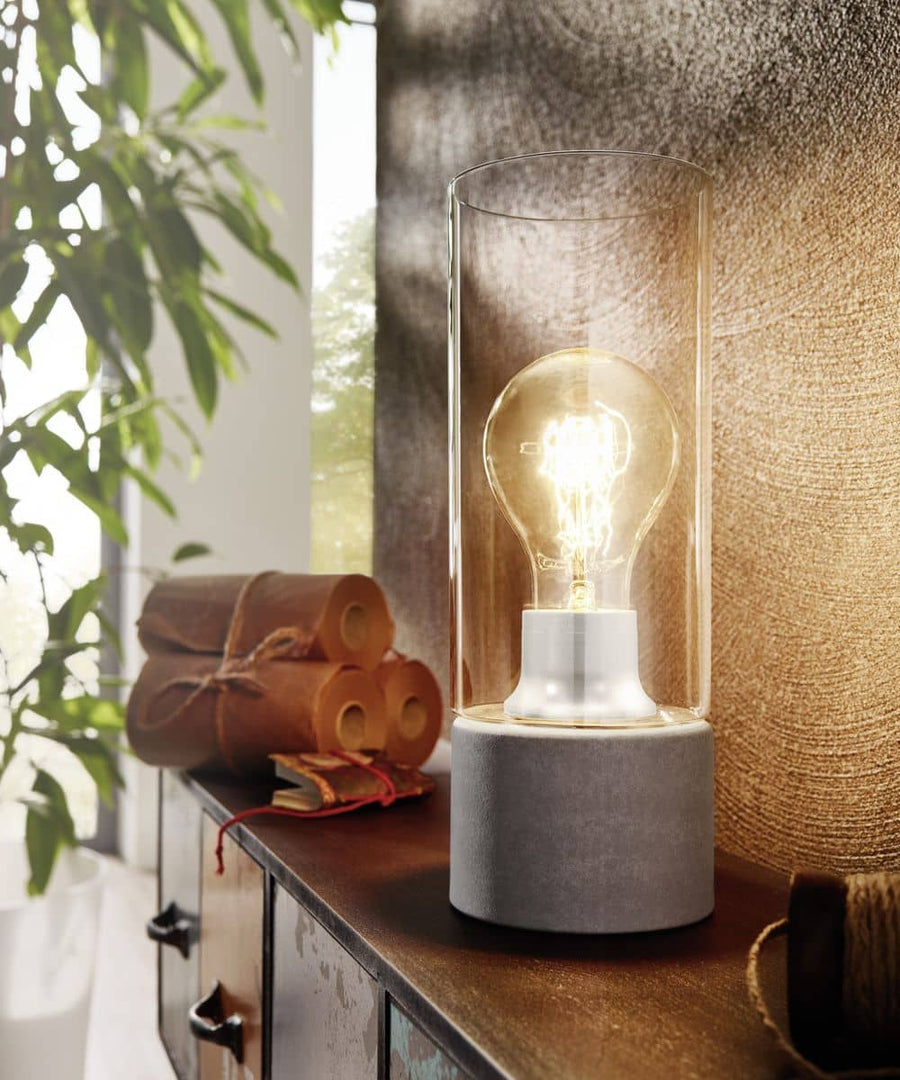 Lampe à poser allumée, socle béton et cylindre de verre, posée sur un meuble