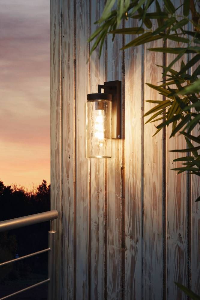 Mise en ambiance extérieure au crépuscule sur bardage bois vertical, applique allumée près d'un feuillage.