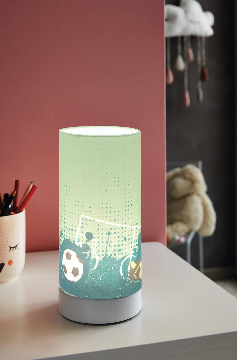 Mise en ambiance de la lampe allumée sur un bureau d'enfant, mur rose et peluches en arrière-plan