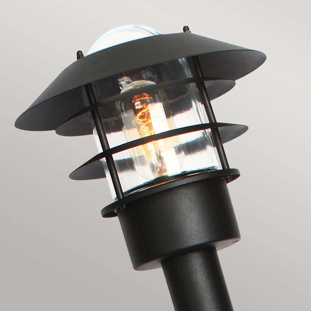 Gros plan sur la tête du lampadaire, ampoule visible derrière une grille métallique noire.