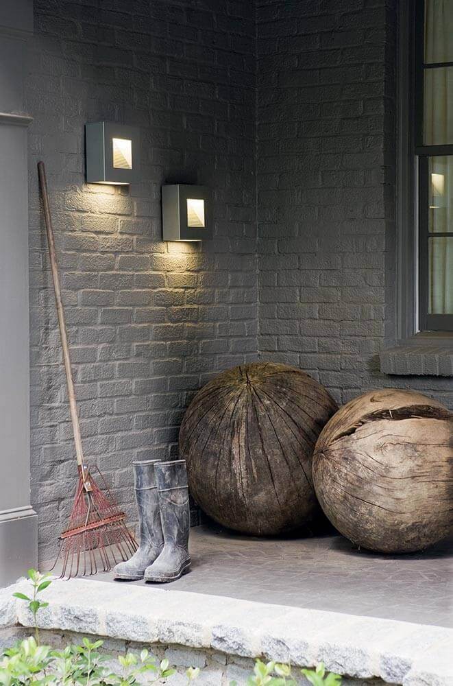Mise en ambiance extérieure avec deux appliques noires sur mur de briques, bottes et râteau.