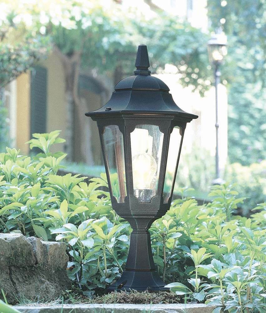 Lampadaire extérieur noir éclairé dans un jardin, entouré de buissons