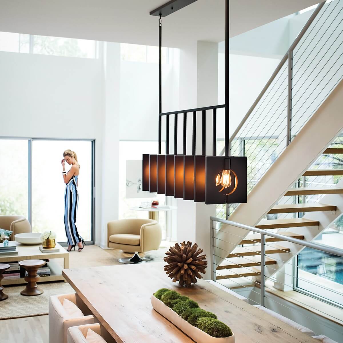 Suspension anthracite installée au-dessus d'une table dans un séjour moderne avec escalier et grande baie vitrée.