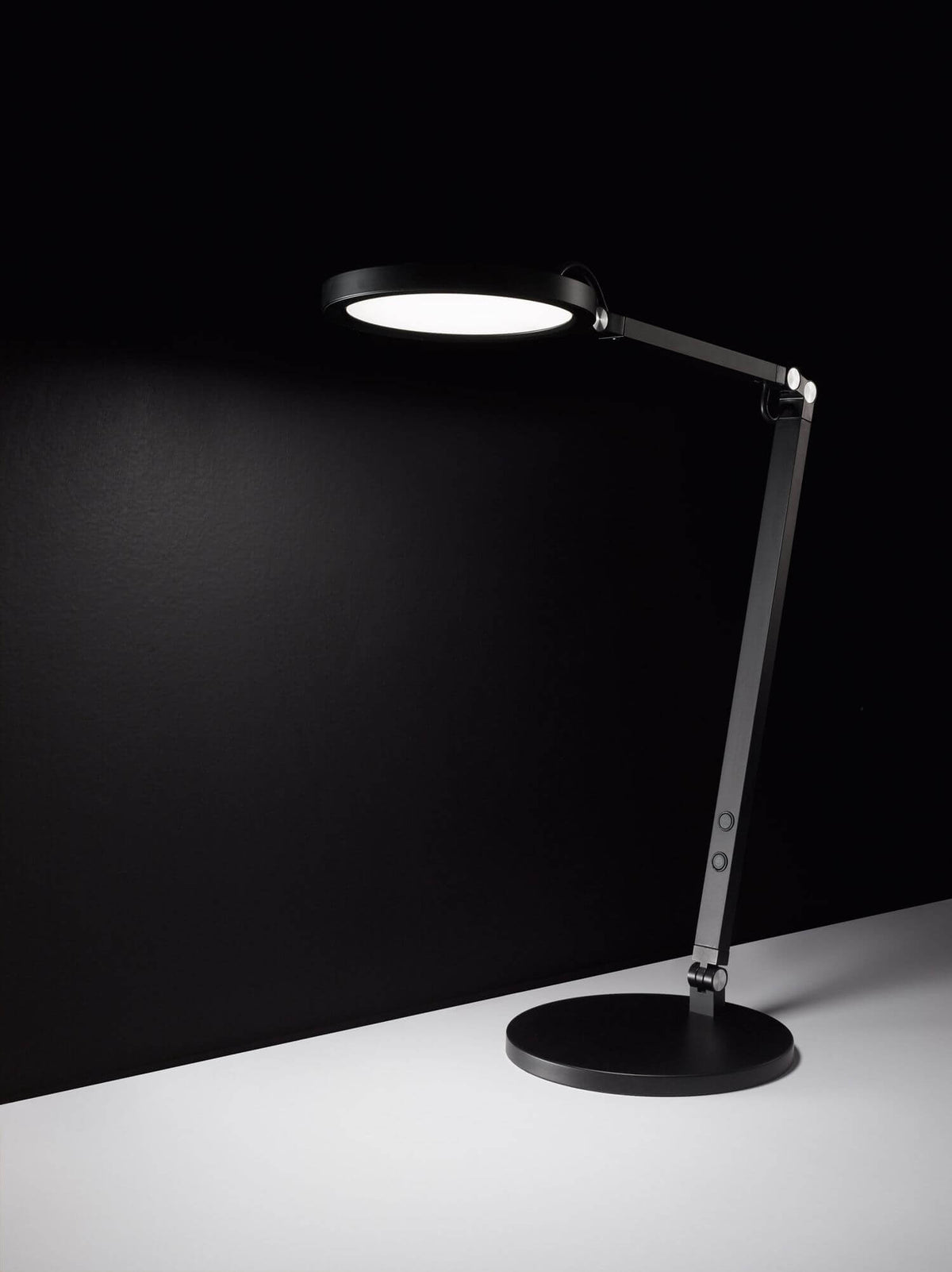 Lampe de bureau noire allumée, abat-jour rond diffusant une lumière blanche sur fond noir.