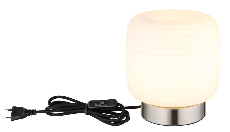 Lampe à poser allumée avec cordon noir, interrupteur et fiche électrique visibles sur fond blanc.
