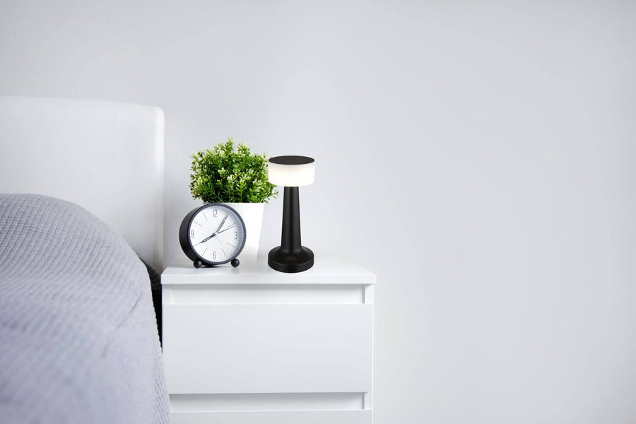Lampe noire allumée posée sur une table de nuit blanche, dans une chambre.