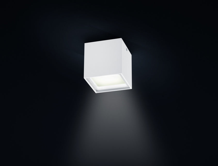Plafonnier blanc cubique allumé, vu de face sur fond sombre.