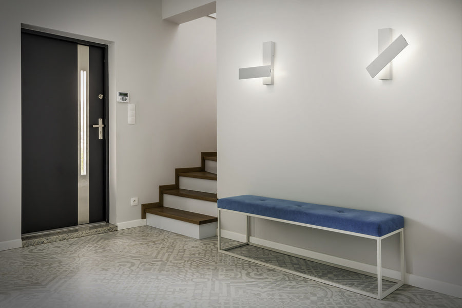 Deux appliques murales allumées dans un couloir clair avec banc bleu et escalier