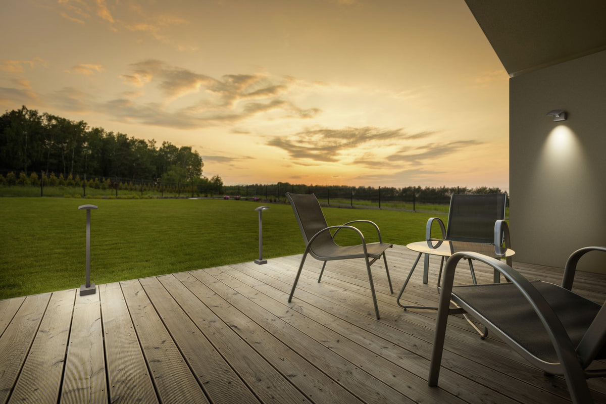 Mise en ambiance sur une terrasse en bois, deux bornes éclairent la pelouse au crépuscule.