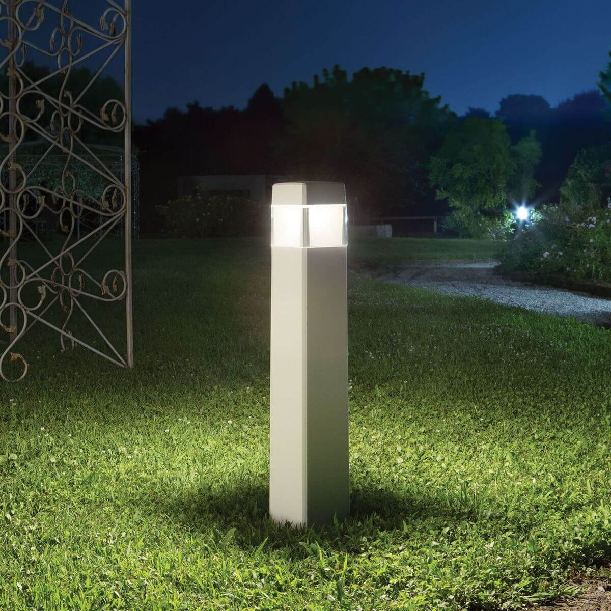 Mise en ambiance nocturne : borne lumineuse blanche éclairée sur une pelouse près d'un portail.