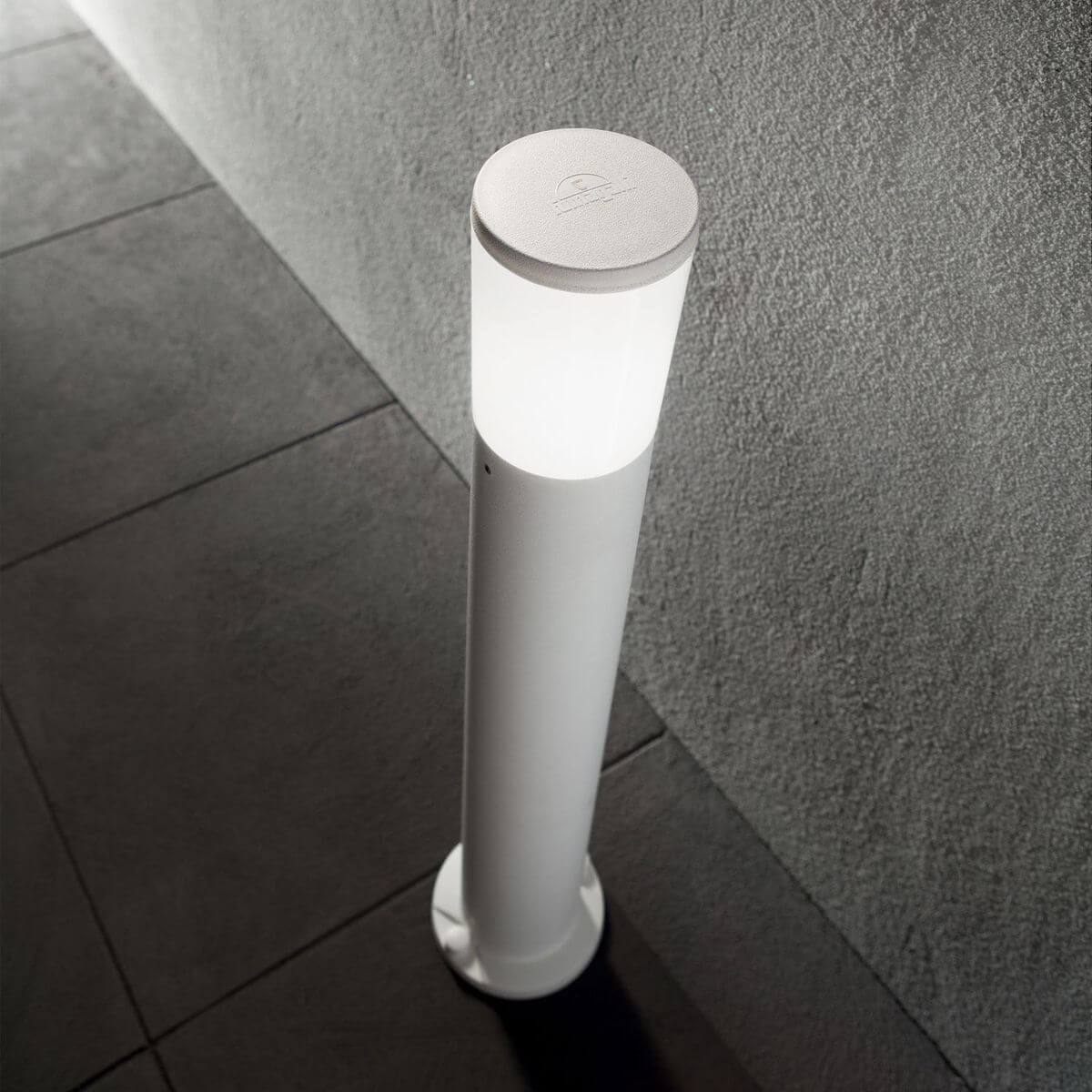 Lampadaire extérieur blanc allumé devant un mur gris, sur des dalles
