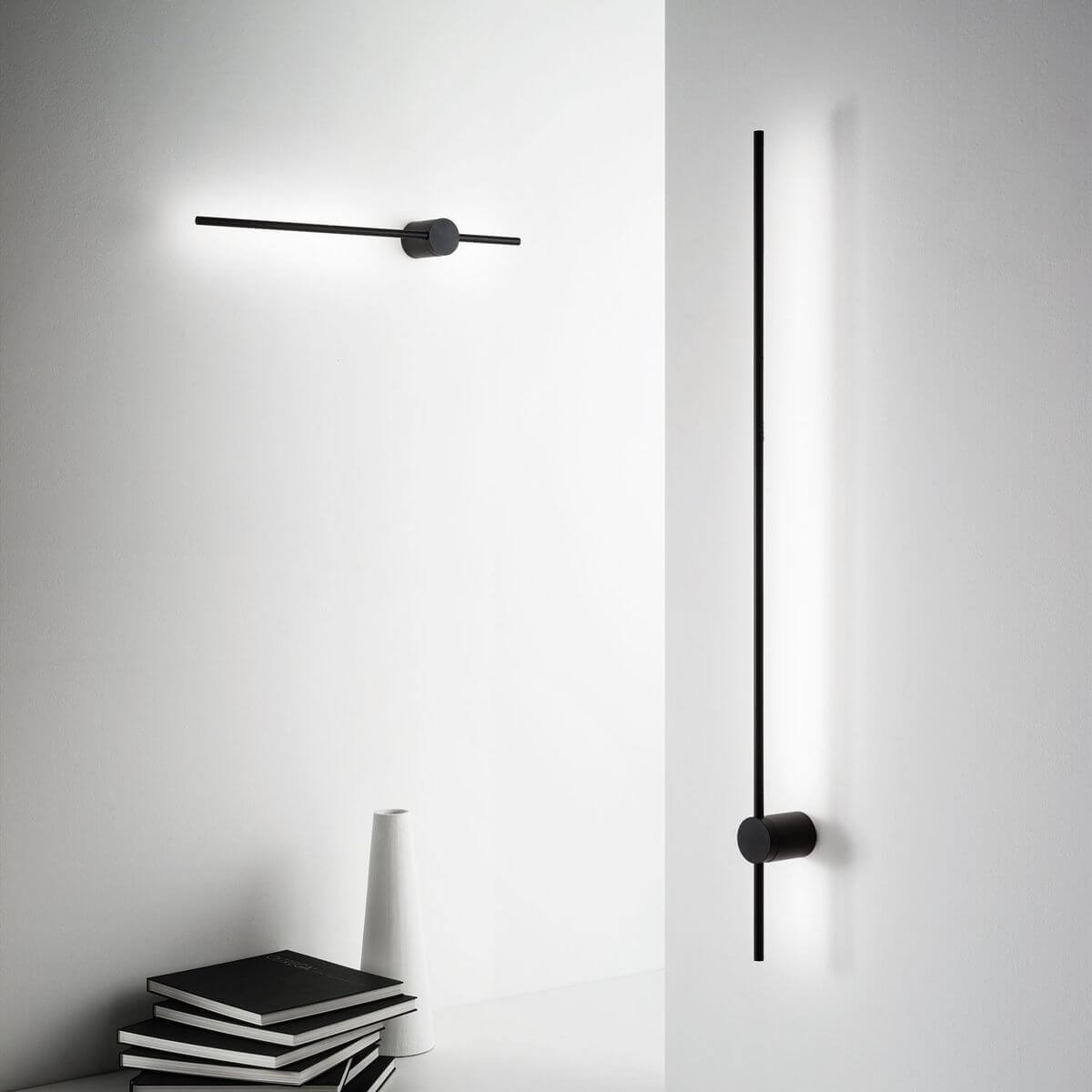 Mise en ambiance murale avec luminaire horizontal et vertical, livres et vase blanc.