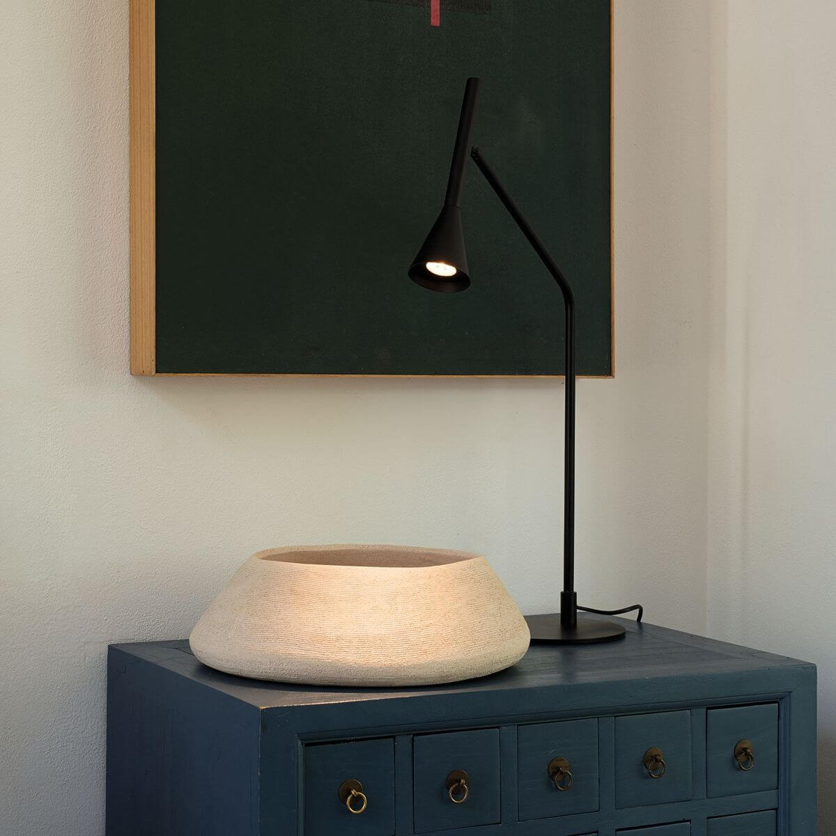 Mise en ambiance de la lampe allumée posée sur une commode bleue devant un tableau