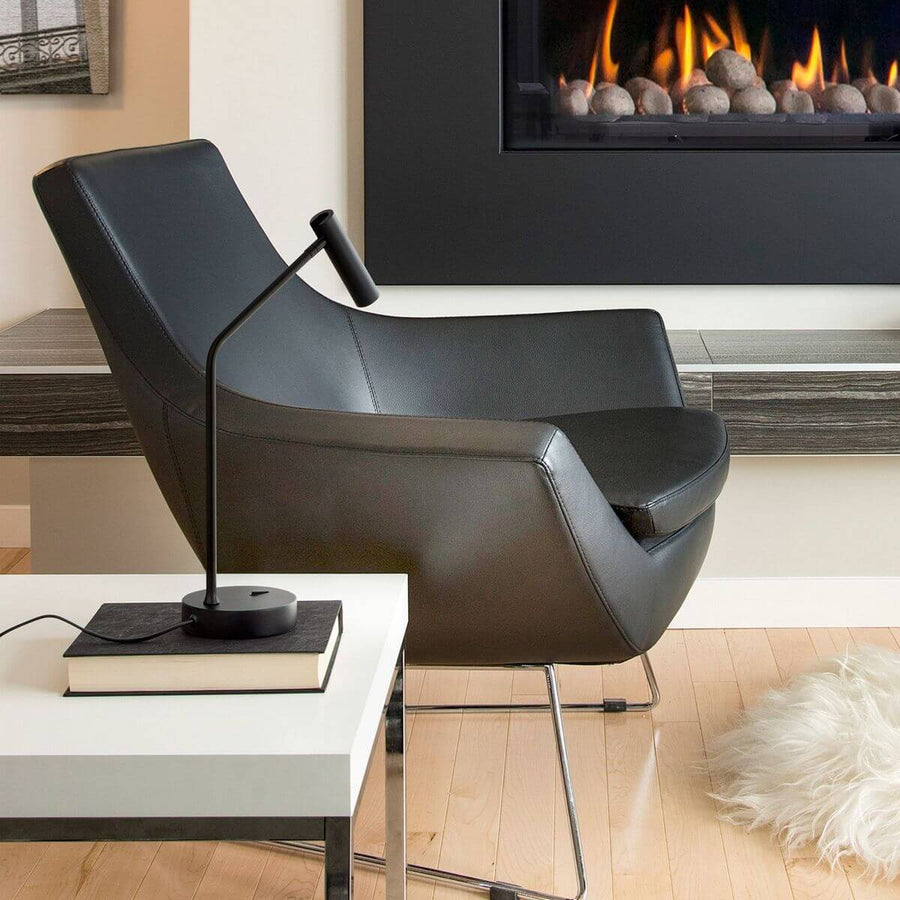 Lampe noire posée sur un livre, près d'un fauteuil en cuir devant une cheminée.