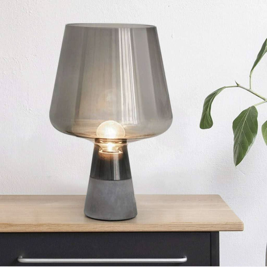 Lampe à poser allumée sur une commode en bois, mur blanc et feuillage à droite.