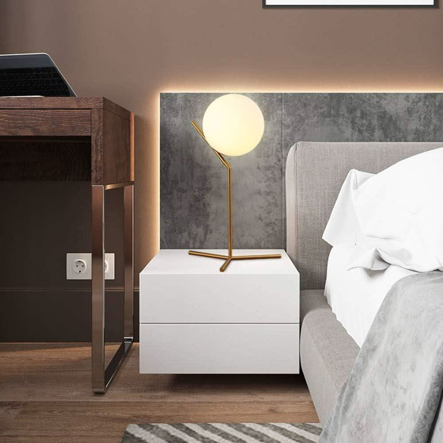 Lampe boule dorée posée sur une table de nuit blanche, tête de lit grise en arrière-plan.