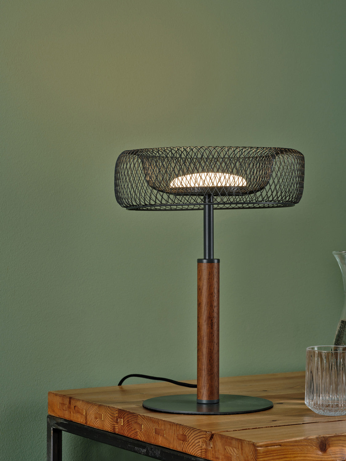 Gros plan de l’abat-jour allumé en maille, pied bois et verre posé sur la table.