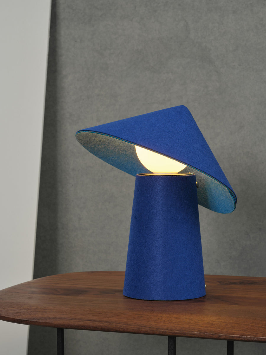 Lampe à poser bleue allumée sur table en bois, abat-jour incliné diffusant une lumière chaude.