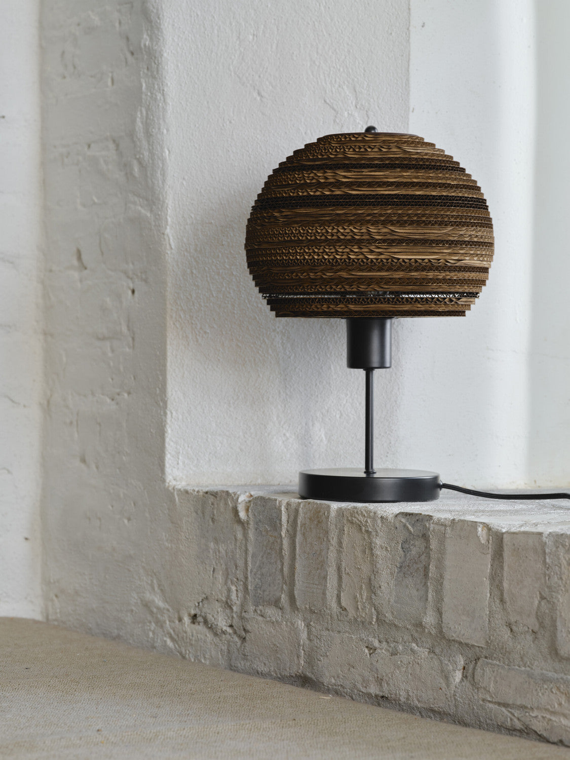 Lampe à poser près d’un mur blanc, pied noir et abat-jour sphérique en carton brun.
