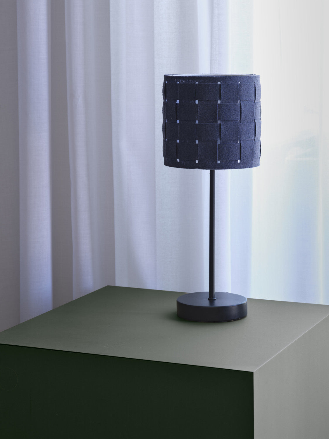 Lampe éteinte devant rideaux blancs, posée sur meuble vert.