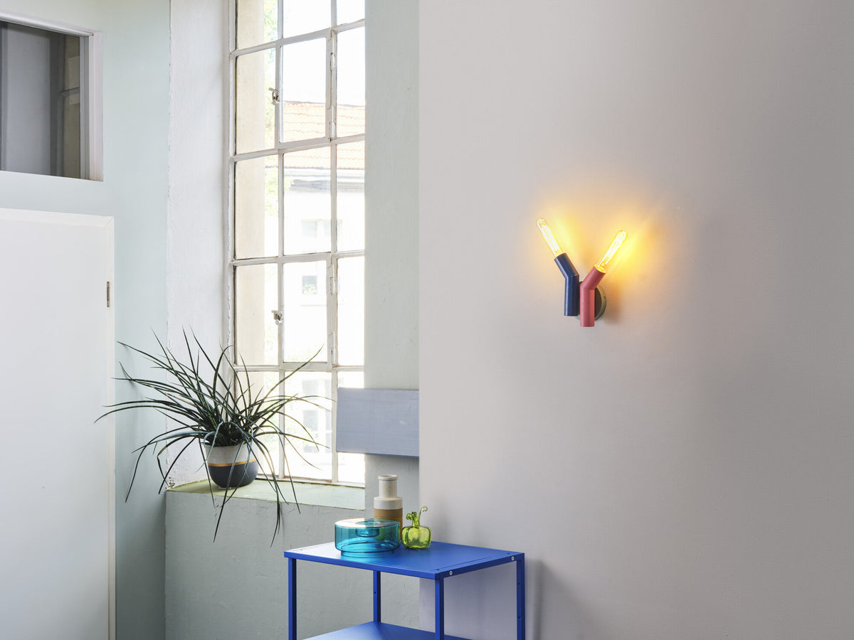 Applique colorée allumée sur un mur blanc, près d'une fenêtre et d'une plante.
