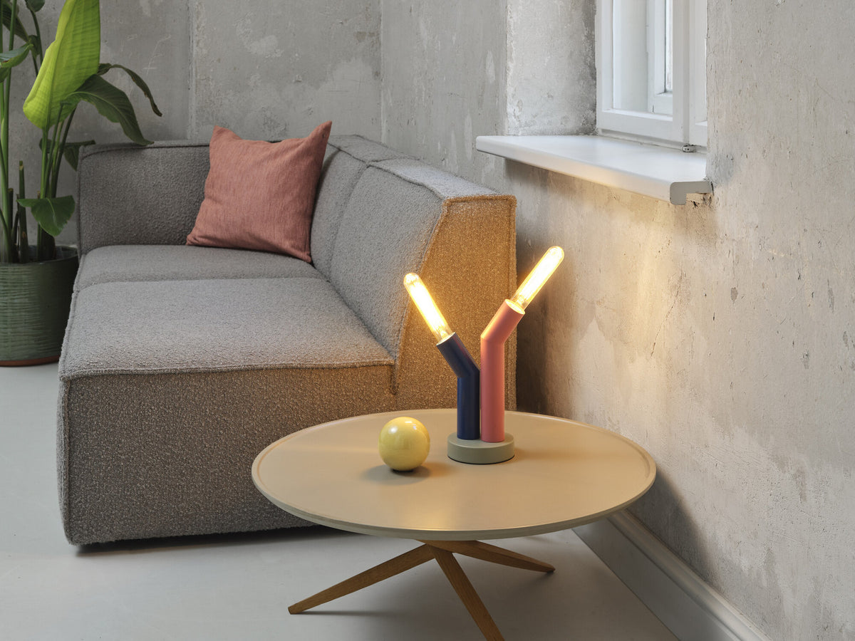 Lampe allumée sur table basse ronde, proche d'un canapé gris et d'un mur béton.
