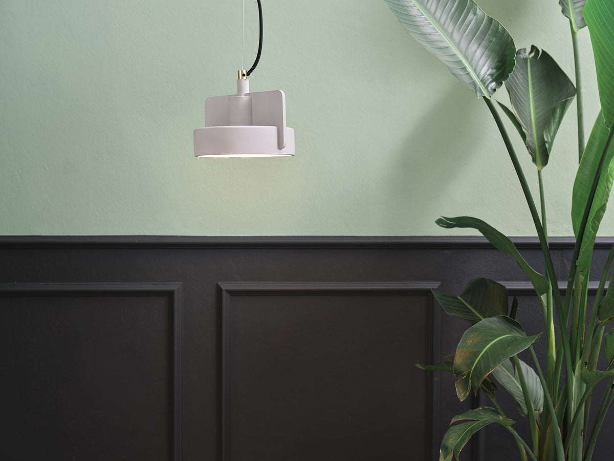 Mise en ambiance : suspension rose pâle devant un mur vert, boiserie noire et grande plante.
