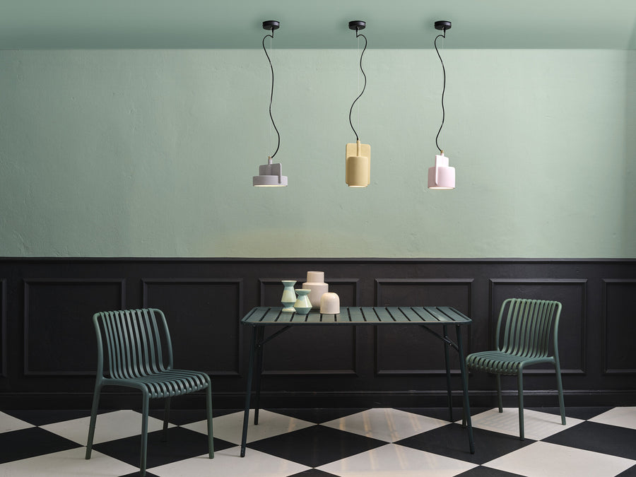 Trois suspensions colorées alignées au-dessus d’une table, chaises vertes et sol en damier.