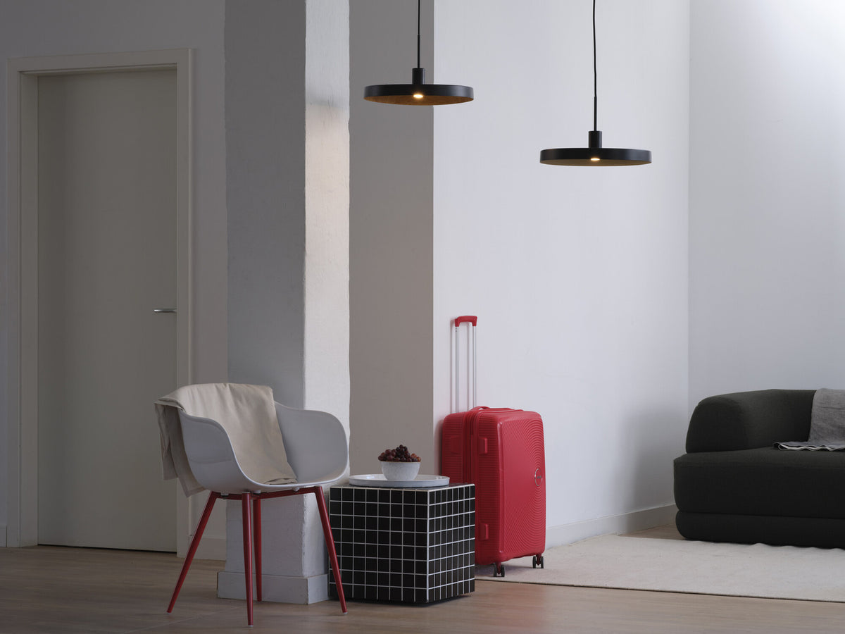 Deux suspensions noires éclairent une pièce avec valise rouge et fauteuil.