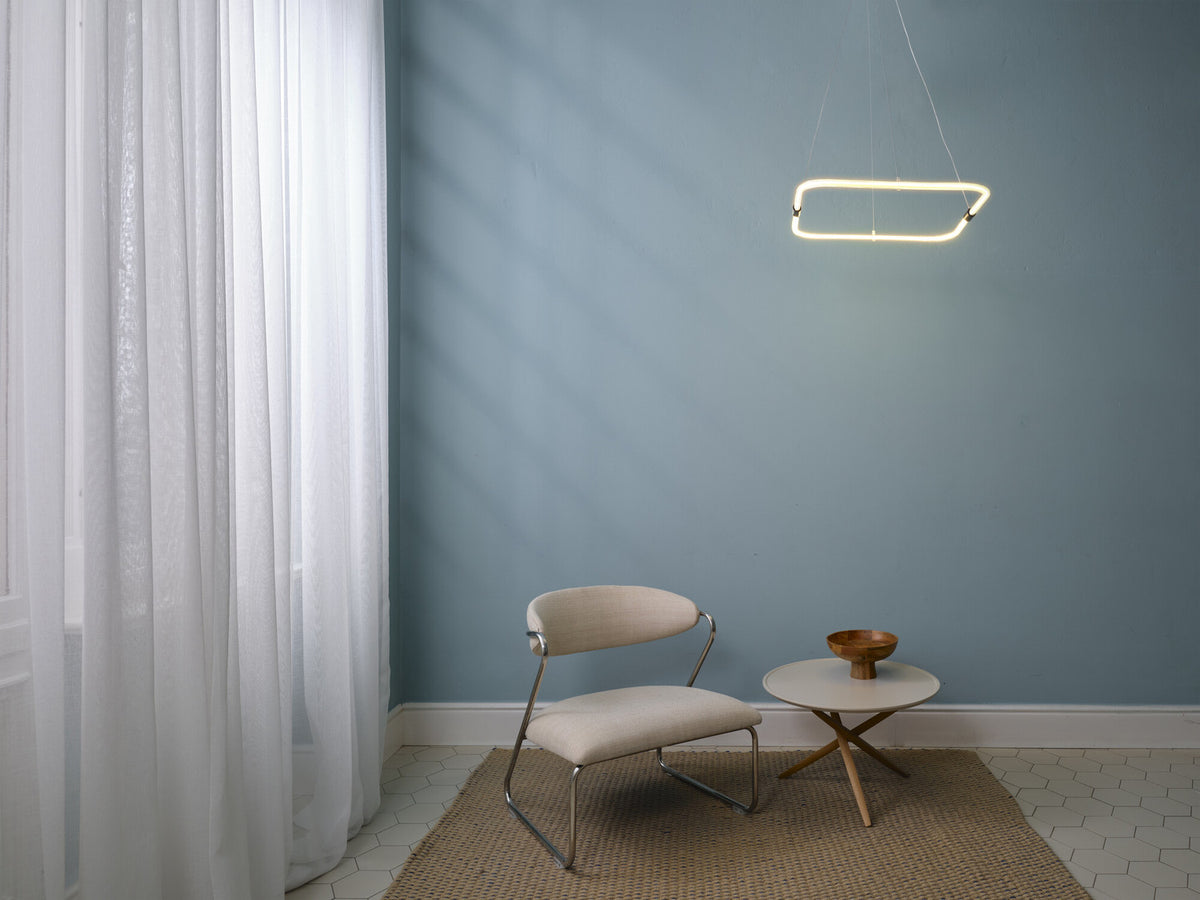 Mise en ambiance dans un salon bleu, suspension rectangulaire allumée au-dessus d’un fauteuil.