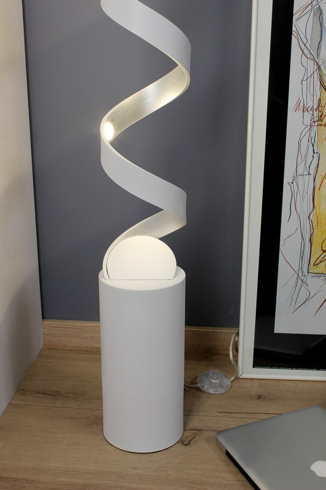 Détail du bas : base cylindrique blanche, spirale lumineuse, câble et sol en bois.