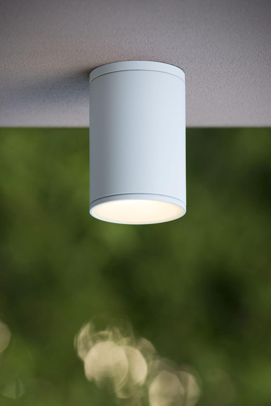 Vue rapprochée du plafonnier blanc cylindrique allumé, installé sous une avancée en extérieur.