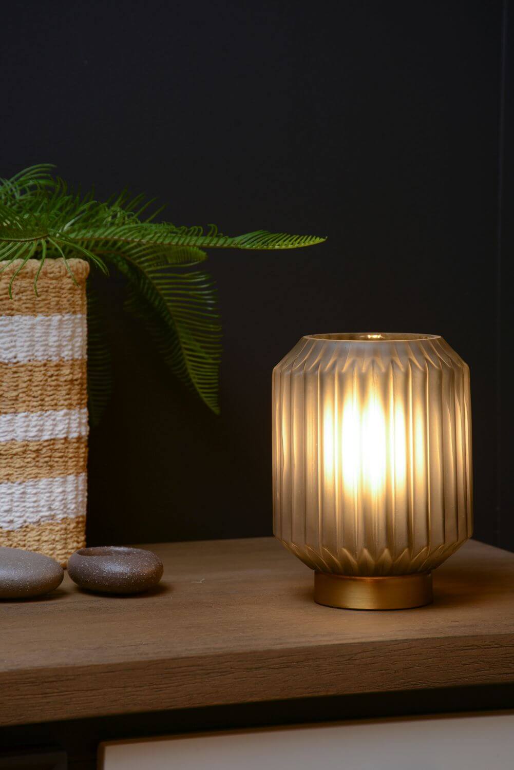 Lampe allumée sur une étagère en bois, près d'un pot tressé et de galets décoratifs.