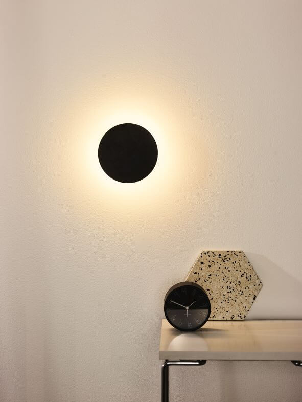 Applique murale ronde noire allumée au-dessus d'une table avec horloge et objet terrazzo
