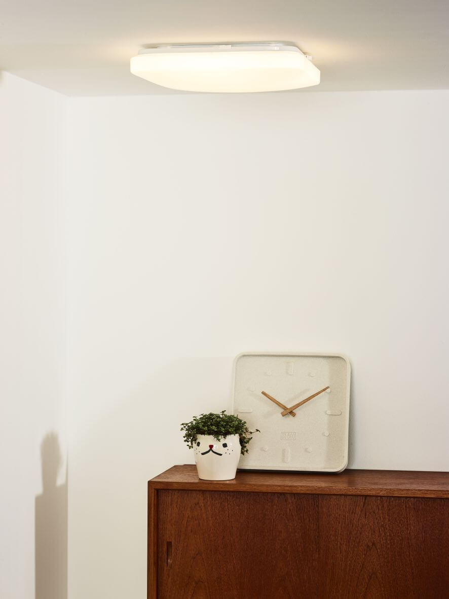 Plafonnier blanc carré allumé au plafond, au-dessus d'un meuble en bois et d'une plante.
