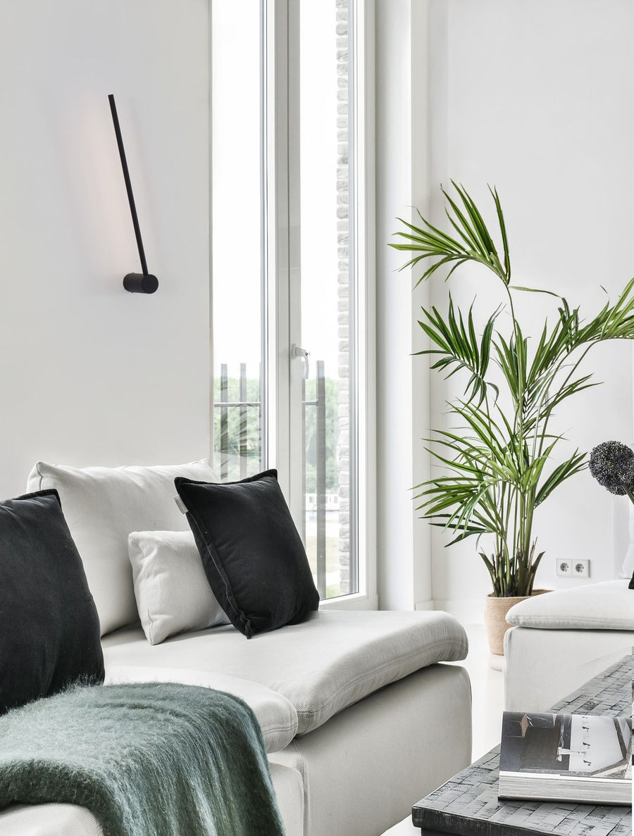 Applique noire fine inclinée sur mur blanc, près d'un canapé et d'une grande fenêtre