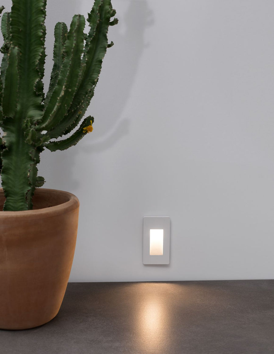 Applique murale blanche allumée sur un mur gris, près d'un cactus en pot