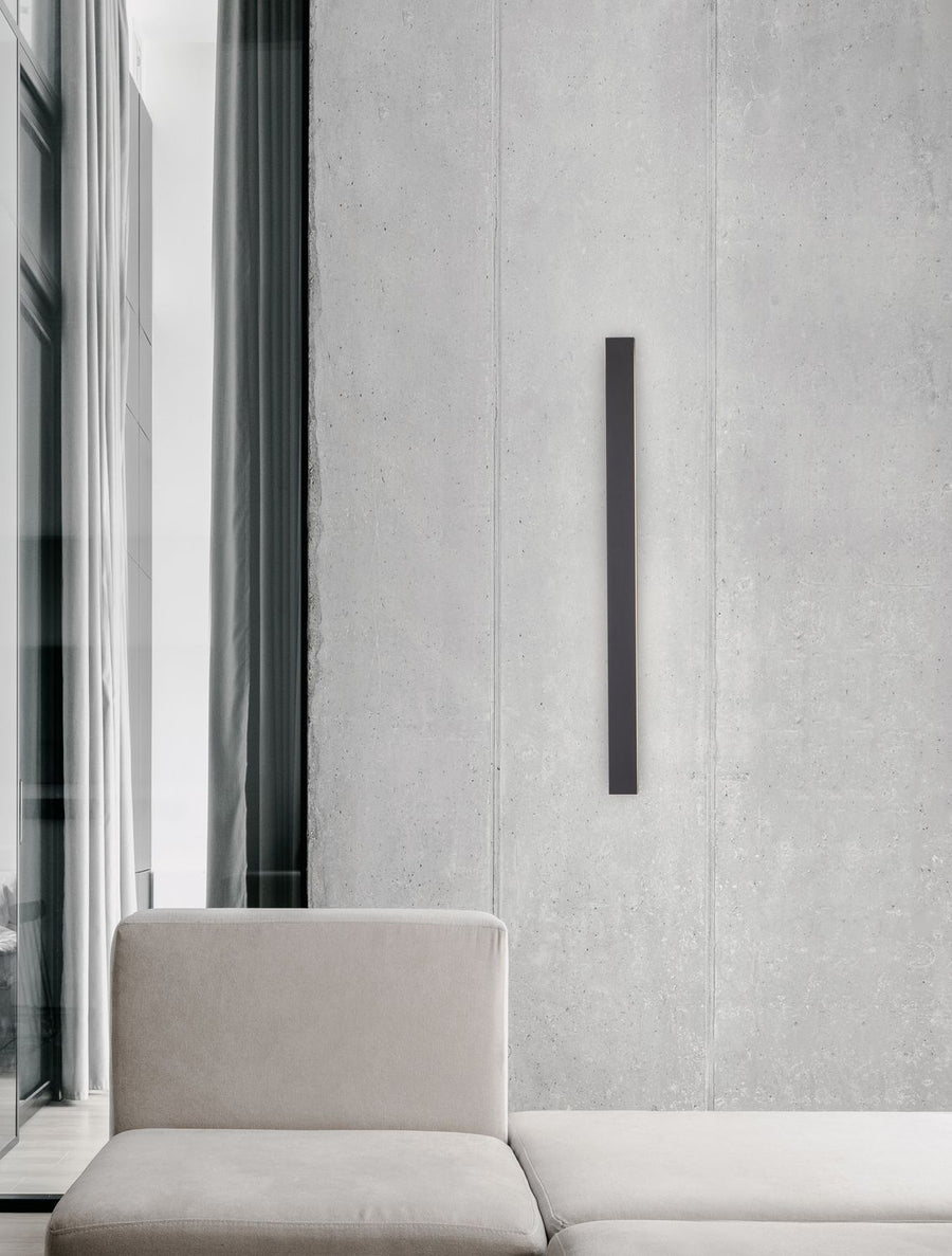 Applique murale verticale noire fixée sur un mur béton, au-dessus d'un canapé beige