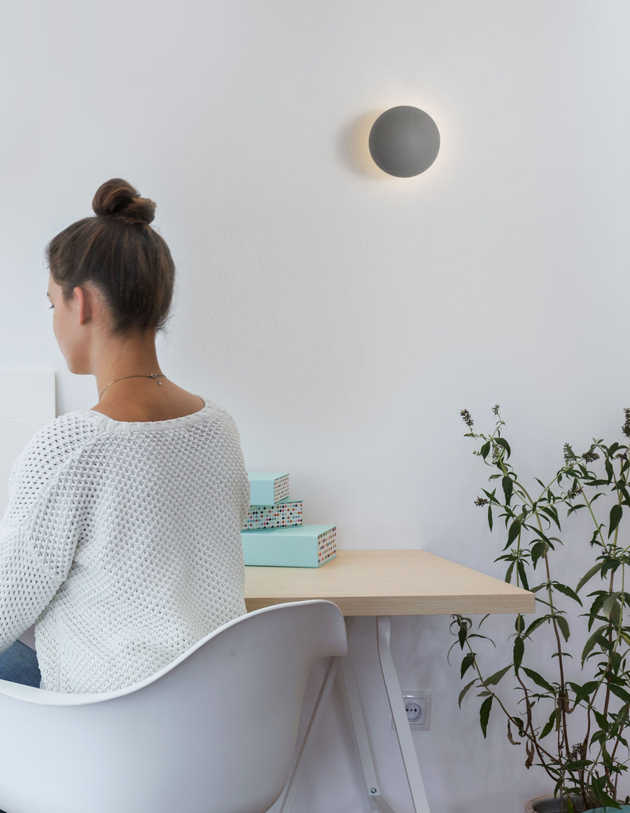 Femme assise à un bureau devant une applique murale grise allumée.