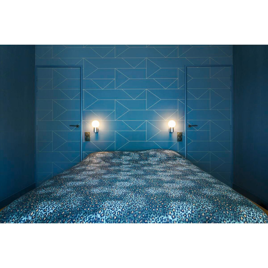 Deux appliques murales éclairent un lit bleu devant un mur géométrique