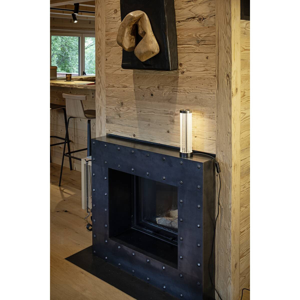 Lampe cylindrique allumée posée sur une cheminée, ambiance chalet en bois clair.