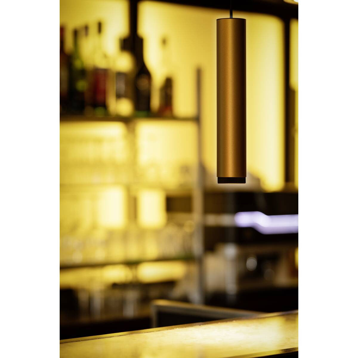 Mise en ambiance au-dessus d’un comptoir de bar, suspension dorée éclairée dans une lumière chaude.