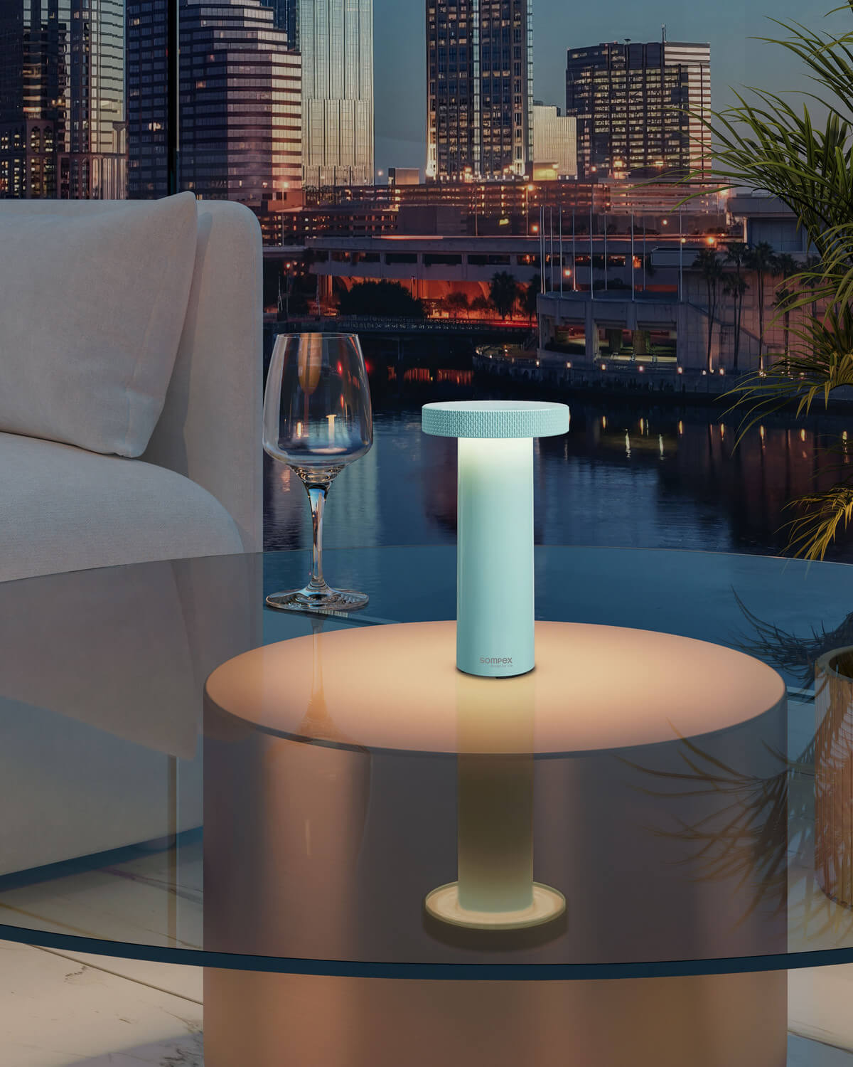 Lampe turquoise allumée posée sur table en verre, vue nocturne urbaine avec canapé et vin.